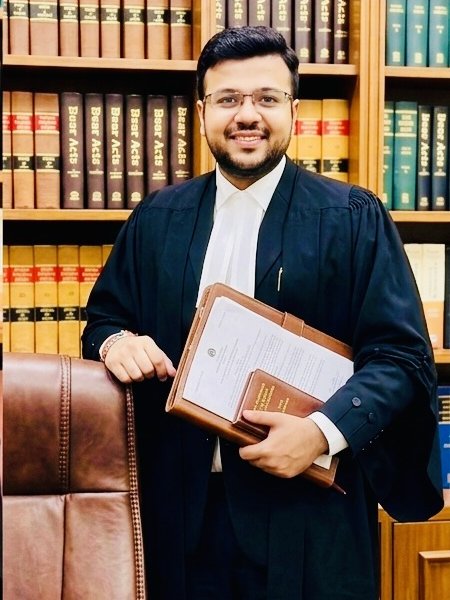Advocate Aayush Agarwal in black advocate robes, standing beside a leather chair in front of law books
