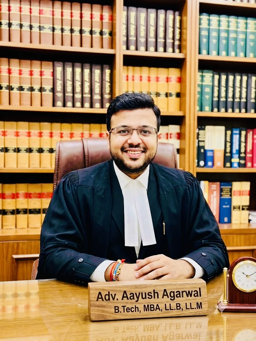 Advocate Aayush Agarwal seated at his desk in black advocate robes, with a nameplate reading B.Tech, MBA, LL.B, LL.M and shelves of law books behind him